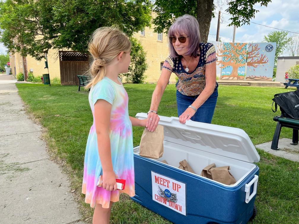 Blonde girl in tie dye dress receives bag lunch handed to her by gray haired woman in a park.