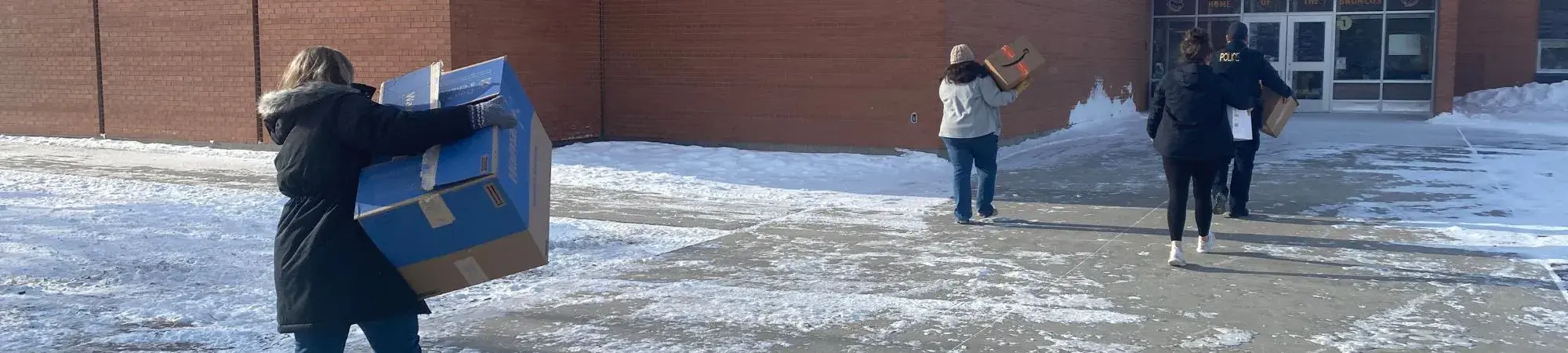 People carry boxes of winter clothing into Falls Elementary school. Snow is on the ground, and people are in heavy coats and boots.