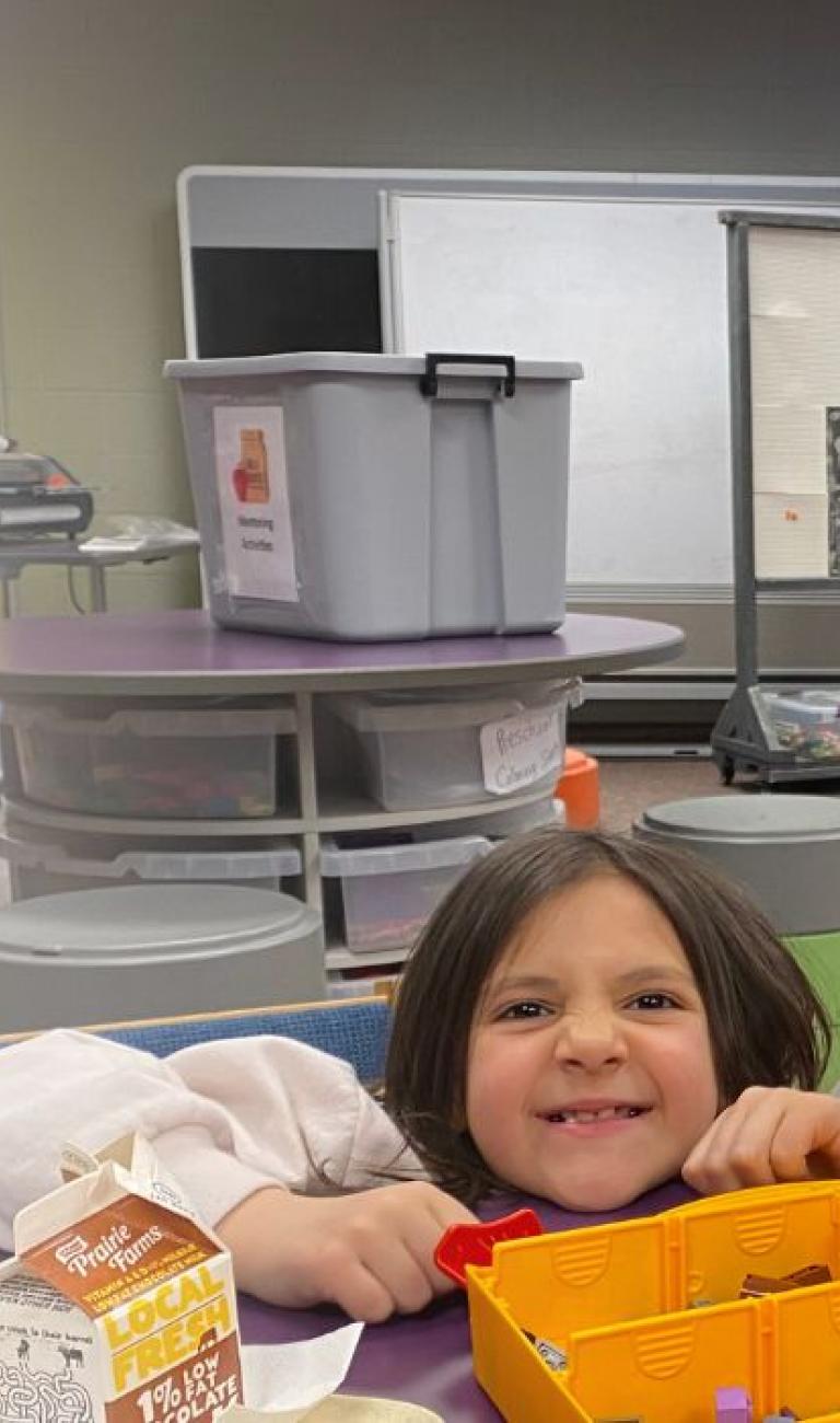 on left, Lunch Buddies logo (brown paper lunch bag with Lunch Buddies mentoring program written on it, an apple nearby). On the right, an image of an elementary student smiling with her mentor.